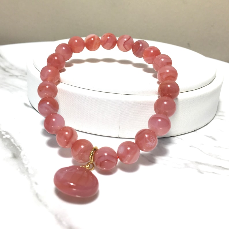May include: A pink agate bracelet with a heart-shaped pendant. The bracelet is made of round beads and has a gold clasp.