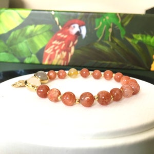 Puede incluir: Un brazalete de piedra de sol con cuentas doradas y un dije dorado. El brazalete est&aacute; hecho de piedras naturales y tiene un color marr&oacute;n claro.