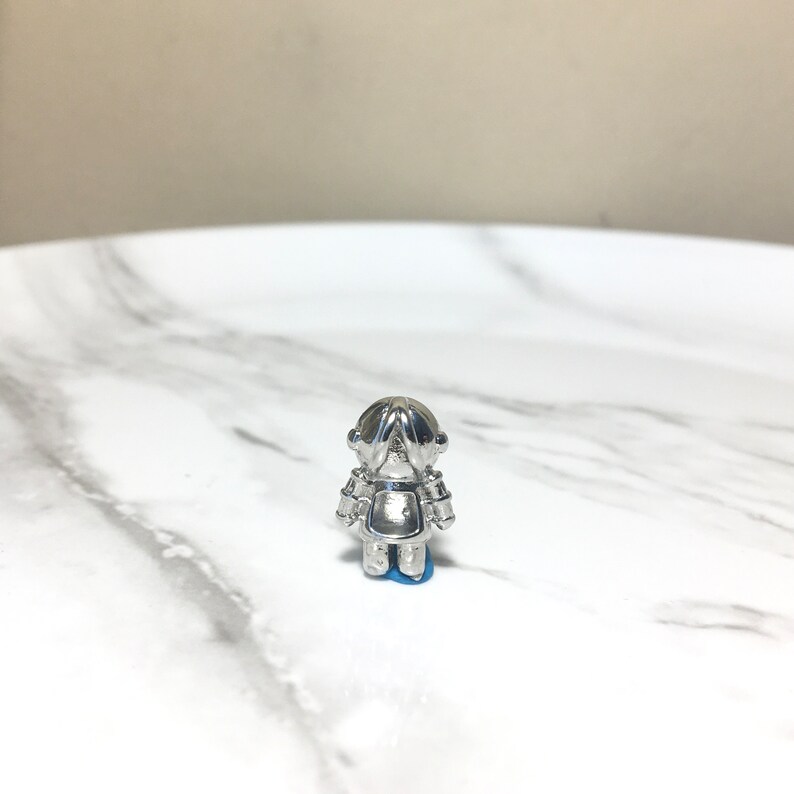 Puede incluir: Una figurita de robot de plata con una base azul. El robot est&aacute; de pie con los brazos cruzados delante de &eacute;l.
