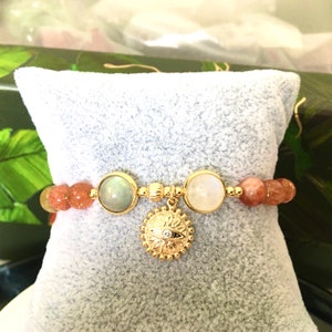 Puede incluir: Un brazalete de oro con un dije de sol y una variedad de cuentas de piedras preciosas, incluyendo piedra de sol, piedra de luna y labradorita.