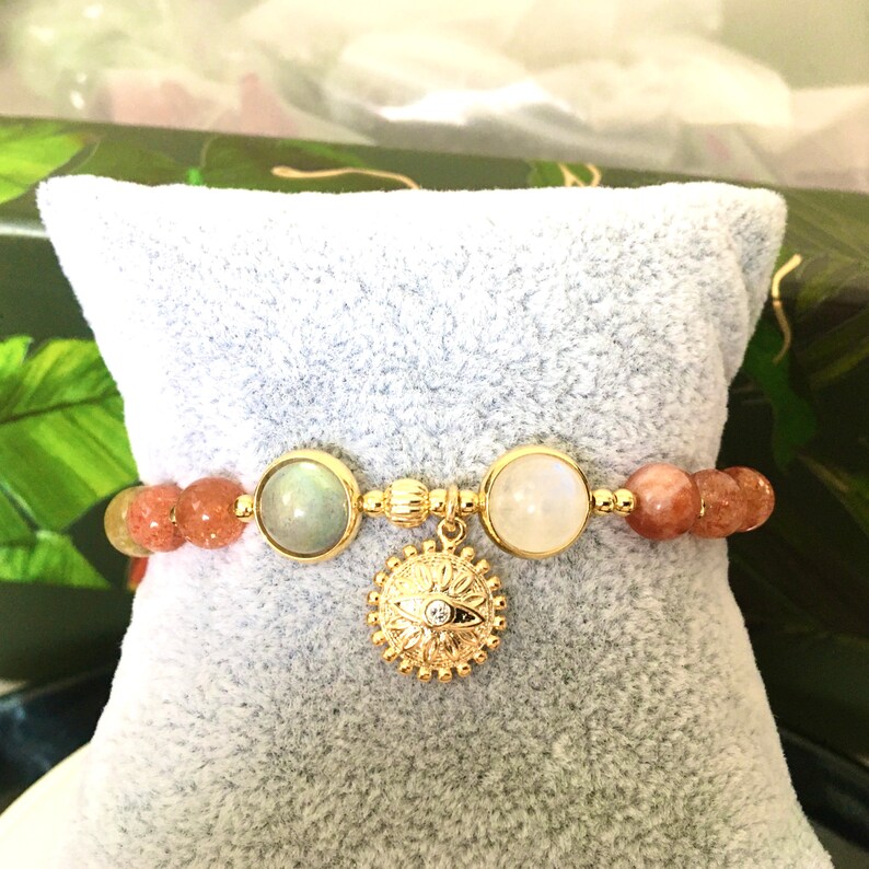Puede incluir: Un brazalete de oro con un dije de sol y una variedad de cuentas de piedras preciosas, incluyendo piedra de sol, piedra de luna y labradorita.