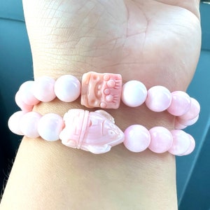 May include: A pink bracelet with two charms, one shaped like a fish and the other like a square with a design. The bracelet is made of round beads.