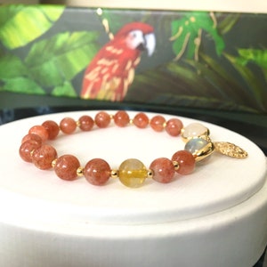 Puede incluir: Un brazalete de oro con cuentas de color marr&oacute;n rojizo y amarillo, una cuenta de piedra lunar blanca y un dije de oro.