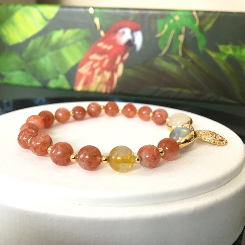 Puede incluir: Un brazalete de oro con cuentas de color marr&oacute;n rojizo y amarillo, una cuenta de piedra lunar blanca y un dije de oro.