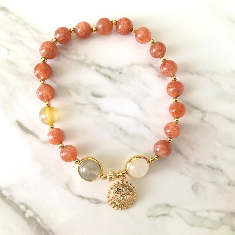 Puede incluir: Un brazalete de oro con un dise&ntilde;o de cuentas de piedra de sol y un dije de oro con una piedra blanca en el centro. El brazalete est&aacute; sobre una superficie de m&aacute;rmol blanco.