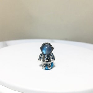 Puede incluir: Un amuleto de astronauta plateado con una piedra de luna azul para la cabeza. El astronauta tiene una estrella en el pecho.