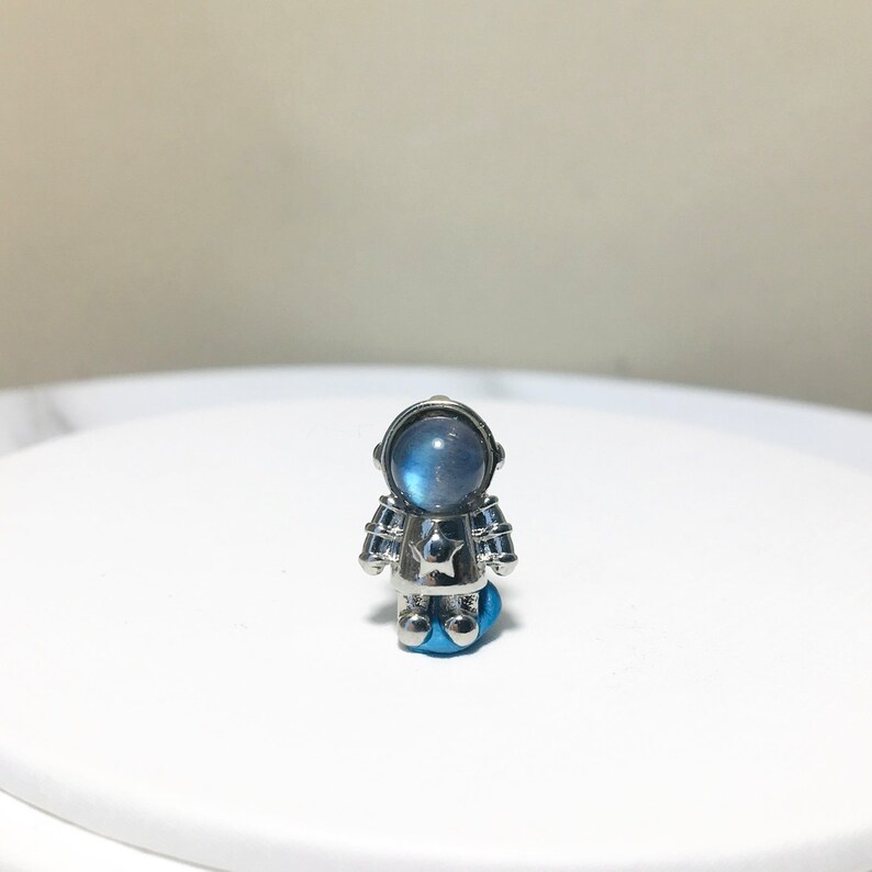 Puede incluir: Un amuleto de astronauta plateado con una piedra de luna azul para la cabeza. El astronauta tiene una estrella en el pecho.