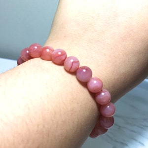 May include: A pink agate beaded bracelet with a natural stone finish. The beads are round and have a light pink colour with white and brown streaks.