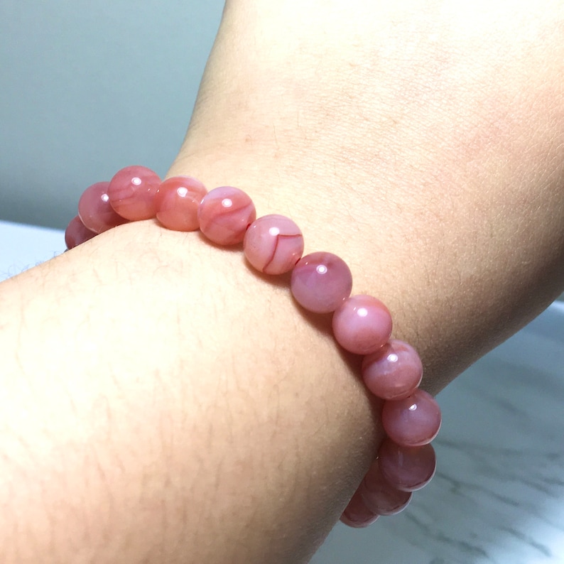 May include: A pink agate beaded bracelet with a natural stone finish. The beads are round and have a light pink colour with white and brown streaks.