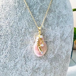 May include: A gold-colored necklace with a large, pink, teardrop-shaped pendant. The pendant is adorned with a gold-colored branch design and three clear, sparkling stones. The necklace is displayed on a gray surface.