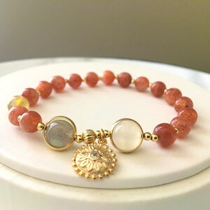 Puede incluir: Un brazalete de oro con cuentas rojas y blancas y un dije de oro con dise&ntilde;o de flor. El brazalete est&aacute; sobre una superficie blanca.