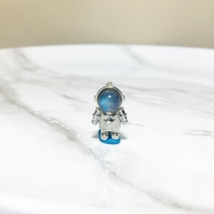 Puede incluir: Un amuleto de astronauta plateado con un cabuj&oacute;n de piedra lunar azul para la cabeza. El astronauta est&aacute; de pie sobre una base azul.