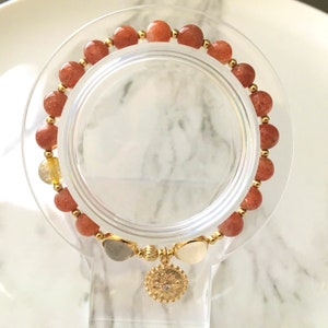 Puede incluir: Un brazalete de oro con un dije de sol y cuentas de piedra de sol rojo-naranja.