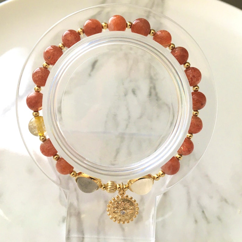 Puede incluir: Un brazalete de oro con un dije de sol y cuentas de piedra de sol rojo-naranja.