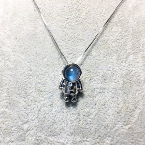 Puede incluir: Un collar de plata con un colgante de astronauta de piedra lunar azul. El astronauta lleva un traje espacial plateado con una estrella en el pecho.