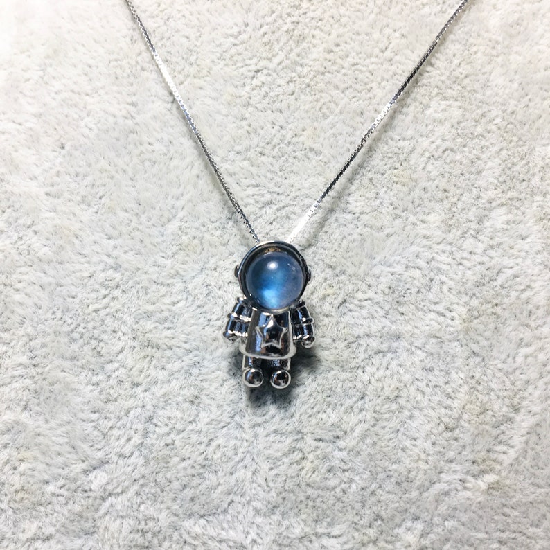 Puede incluir: Un collar de plata con un colgante de astronauta de piedra lunar azul. El astronauta lleva un traje espacial plateado con una estrella en el pecho.
