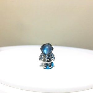 Puede incluir: Un amuleto de astronauta plateado con una cara de piedra lunar azul y una estrella en el pecho.