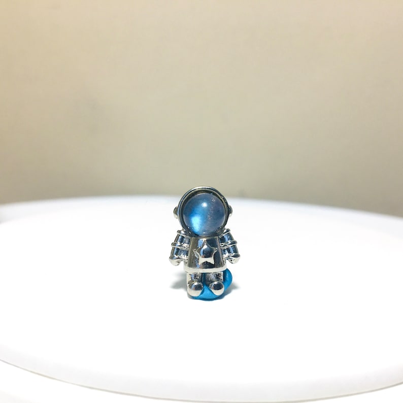 Puede incluir: Un amuleto de astronauta plateado con una cara de piedra lunar azul y una estrella en el pecho.