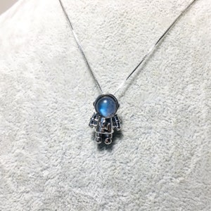 Puede incluir: Un collar de plata con un colgante de astronauta de piedra lunar. El astronauta lleva un traje espacial plateado con una piedra lunar azul para la cabeza.