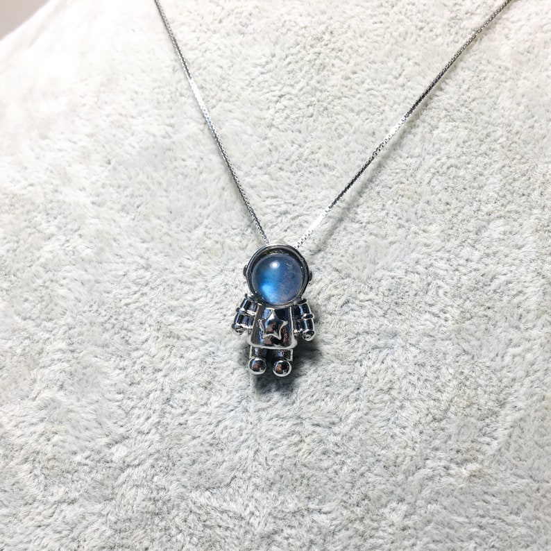Puede incluir: Un collar de plata con un colgante de astronauta de piedra lunar. El astronauta lleva un traje espacial plateado con una piedra lunar azul para la cabeza.