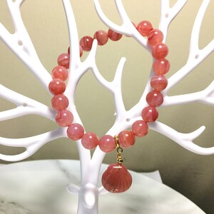 May include: A pink agate beaded bracelet with a pink agate seashell charm. The bracelet is displayed on a white branch-like stand.