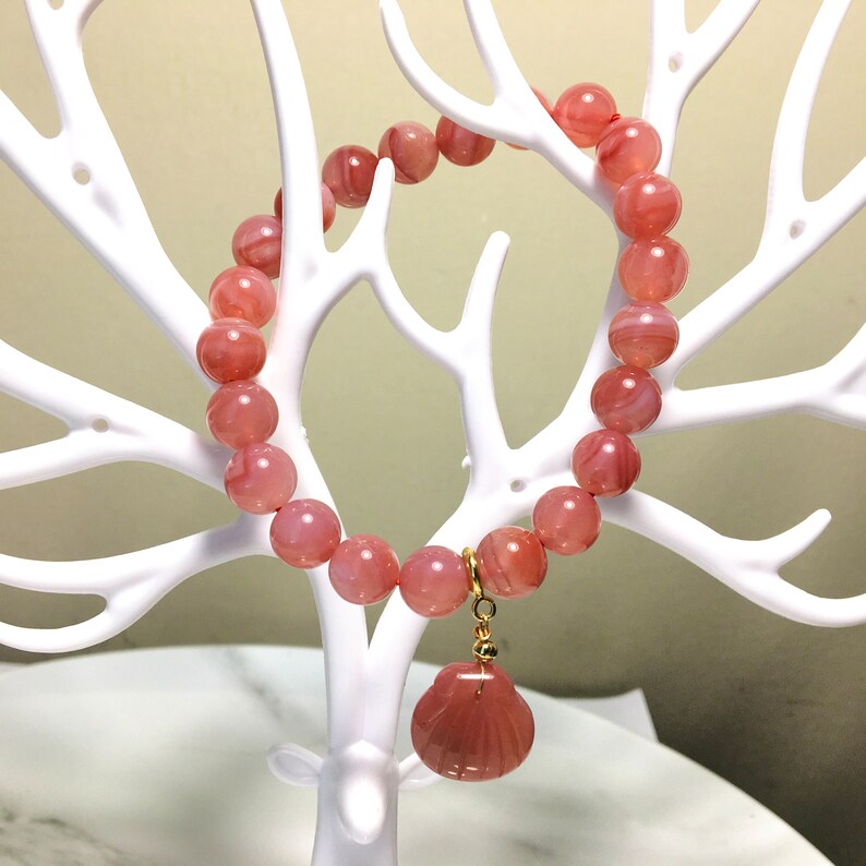 May include: A pink agate beaded bracelet with a pink agate seashell charm. The bracelet is displayed on a white branch-like stand.