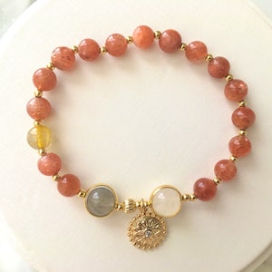 Puede incluir: Un brazalete de oro con un dise&ntilde;o de cuentas de piedra de sol y un dije de oro con un patr&oacute;n floral. El brazalete tiene una mezcla de cuentas de color marr&oacute;n claro y marr&oacute;n oscuro.
