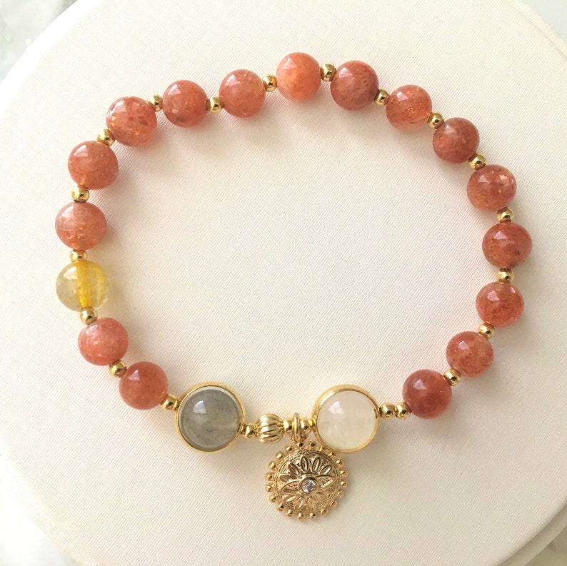 Puede incluir: Un brazalete de oro con un dise&ntilde;o de cuentas de piedra de sol y un dije de oro con un patr&oacute;n floral. El brazalete tiene una mezcla de cuentas de color marr&oacute;n claro y marr&oacute;n oscuro.