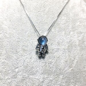 Puede incluir: Un collar de plata con un peque&ntilde;o colgante de astronauta. La cabeza del astronauta es una piedra preciosa azul.