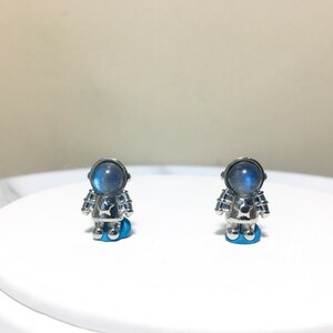 Puede incluir: Un par de pendientes de astronauta de plata con cabujones de piedra lunar azul. Los pendientes est&aacute;n sobre una superficie blanca.