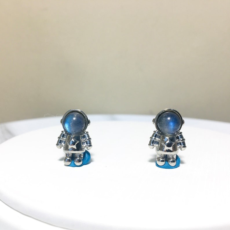 Puede incluir: Un par de pendientes de astronauta de plata con cabujones de piedra lunar azul. Los pendientes est&aacute;n sobre una superficie blanca.