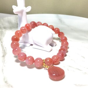 May include: A pink agate gemstone bracelet with a pink agate pendant. The bracelet is made of round beads and has a gold clasp.