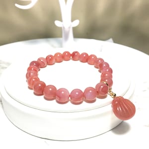 May include: A pink agate gemstone bracelet with a pink agate shell charm. The bracelet is made of round beads and has a gold clasp.