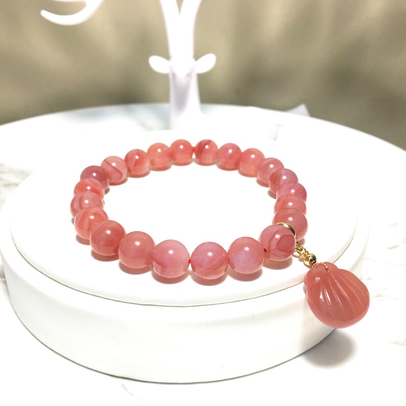 May include: A pink agate gemstone bracelet with a pink agate shell charm. The bracelet is made of round beads and has a gold clasp.