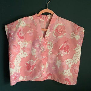 May include: A pink floral print top with a V-neck and short sleeves. The top has a button-down front and is hanging on a pink hanger. The floral pattern includes shades of pink and white.