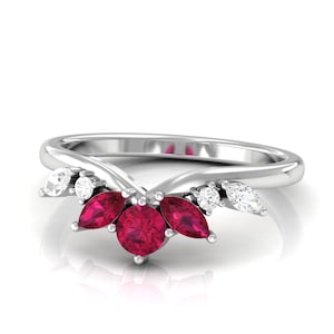 May include: A silver ring featuring a cluster of three round, deep red gemstones at the center, flanked by marquise and round white gemstones. The ring's band curves upward, creating a delicate, elegant design.