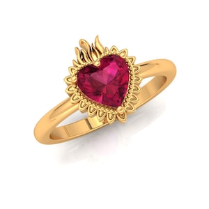 May include: A gold-colored ring featuring a heart-shaped, ruby-colored gemstone. The gemstone is surrounded by a sunburst design with a flame detail at the top. The ring band is smooth and polished.
