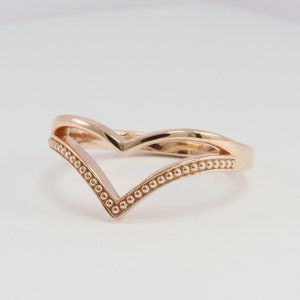 May include: A rose gold ring with a V-shaped design. The ring is accented with a beaded edge.