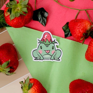 Puede incluir: Una pegatina de una rana verde claro con un sombrero de fresa, con la lengua fuera. La pegatina está rodeada de fresas rojas frescas y hojas verdes, sobre un fondo verde y rosa.