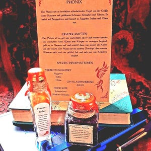 Könnte beinhalten: Ein Bild mit einer Sammlung von Gegenständen, die sich auf den mythischen Phönix beziehen. Es zeigt eine kleine Glasflasche mit der Aufschrift "Phönixfeder", ein Glas "Phönixasche" und eine dekorative Karte mit Text und Illustrationen. Die Gegenstände sind mit Büchern und Stiften angeordnet.