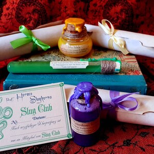 Könnte beinhalten: Eine grüne Schlangenillustration auf einer Karte mit dem Text "Prof. Horace Slughorns Slug Club" und "Nr. Datum: Wach auffälliger & Serprüfung ist ein Mitglied vom Slug Club". Es gibt drei kleine Flaschen mit lila, gelber und grüner Flüssigkeit, jede mit einem Korkstopfen und einem Wachssiegel. Zwei aufgerollte Schriftrollen mit Bändern sind ebenfalls im Bild.