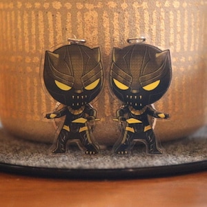Peut inclure: Deux breloques de personnages noirs et dorés de Black Panther, un super-héros Marvel. Les breloques se font face et sont sur une surface grise.