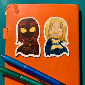 May include: Two cartoon character stickers on an orange background. The sticker on the left is a dark figure with yellow eyes and a red cape. The sticker on the right is a blonde character with a yellow suit and blue cape.