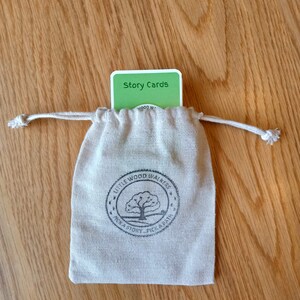 May include: A small, beige drawstring bag with a printed logo and a stack of green "Story Cards" peeking out. The bag features a tree logo and the text "Little Wood Walkers". The cards have white text on a green background.