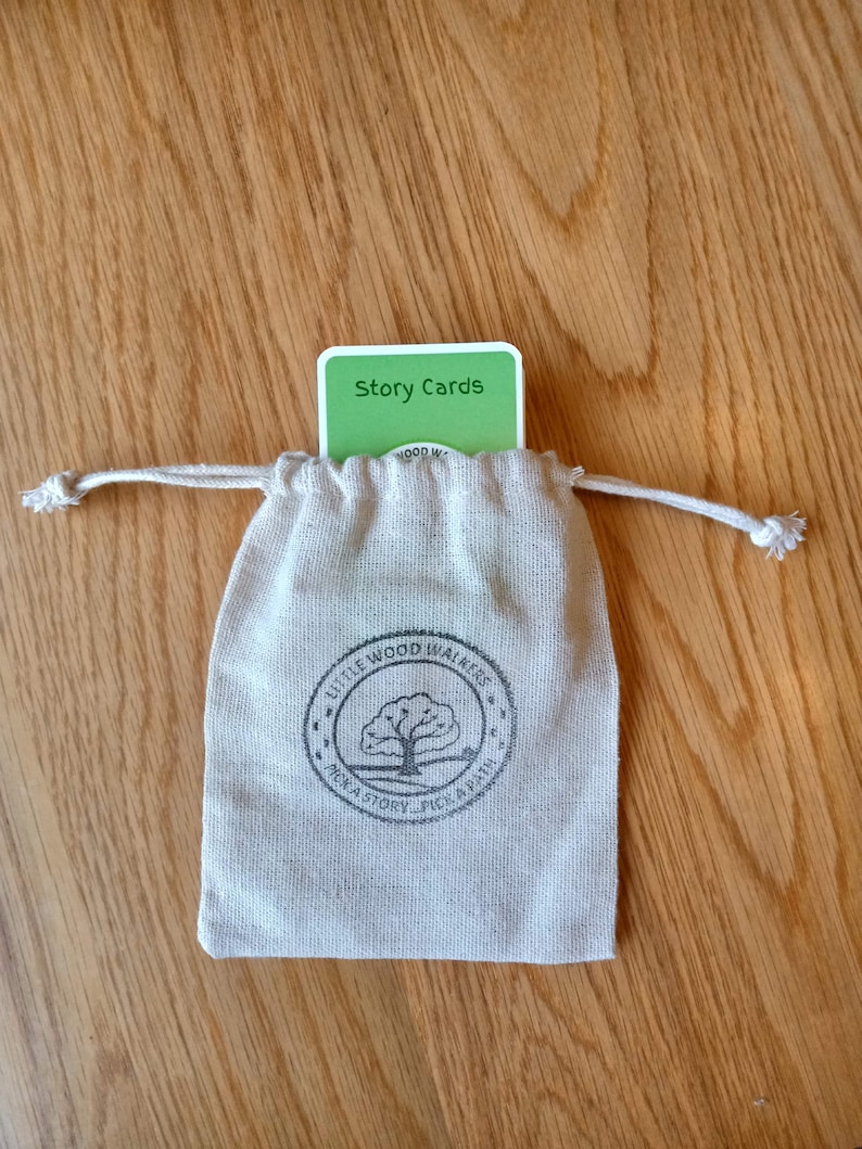 May include: A small, beige drawstring bag with a printed logo and a stack of green "Story Cards" peeking out. The bag features a tree logo and the text "Little Wood Walkers". The cards have white text on a green background.