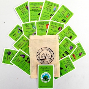 May include: A set of green cards with illustrated recipes for mud kitchen creations. The cards are arranged in a circle around a small, white canvas bag with a brown logo that reads "Little Wood Walkers Pick a Story Pick a Path".