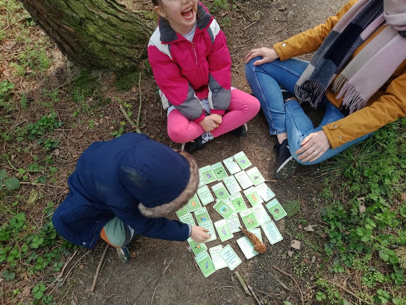 May include: A group of cards with illustrations and text are laid out on the ground. A child in a blue jacket and another in a pink jacket are interacting with the cards. An adult in a yellow jacket and blue jeans is also present.