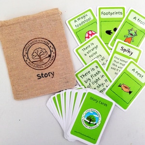 May include: A set of "Story Cards" with a burlap bag. The cards are green and white, featuring illustrations and text prompts like "Footprints" and "A troll." The bag is printed with "Little Wood Walkers" and "Pick a Story, Pick a Path."