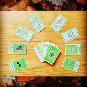 May include: A collection of green and white story cards with illustrations and text, including "A dragon," "Footprints," and "Scared." The cards are arranged on a wooden surface, with a deck of cards in the center.