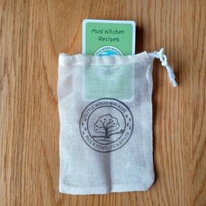 May include: A white drawstring bag with a green card inside. The card has the text "Mud Kitchen Recipes" and the logo "Little Wood Walkers Pick a Story... Pick a Path".